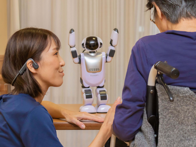 白と黒のロボットが両手を上げている。左側には青い服を着た人物、右側には車椅子に座った人物がいる。